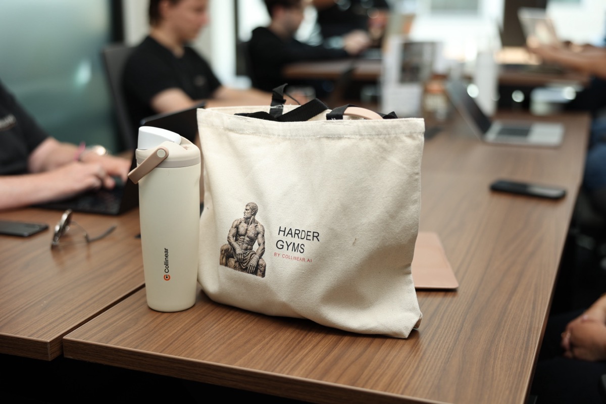 A canvas tote bag printed with 'Harder Gyms by Collinear.ai' and a stone-statue illustration, resting on a desk next to a water bottle.