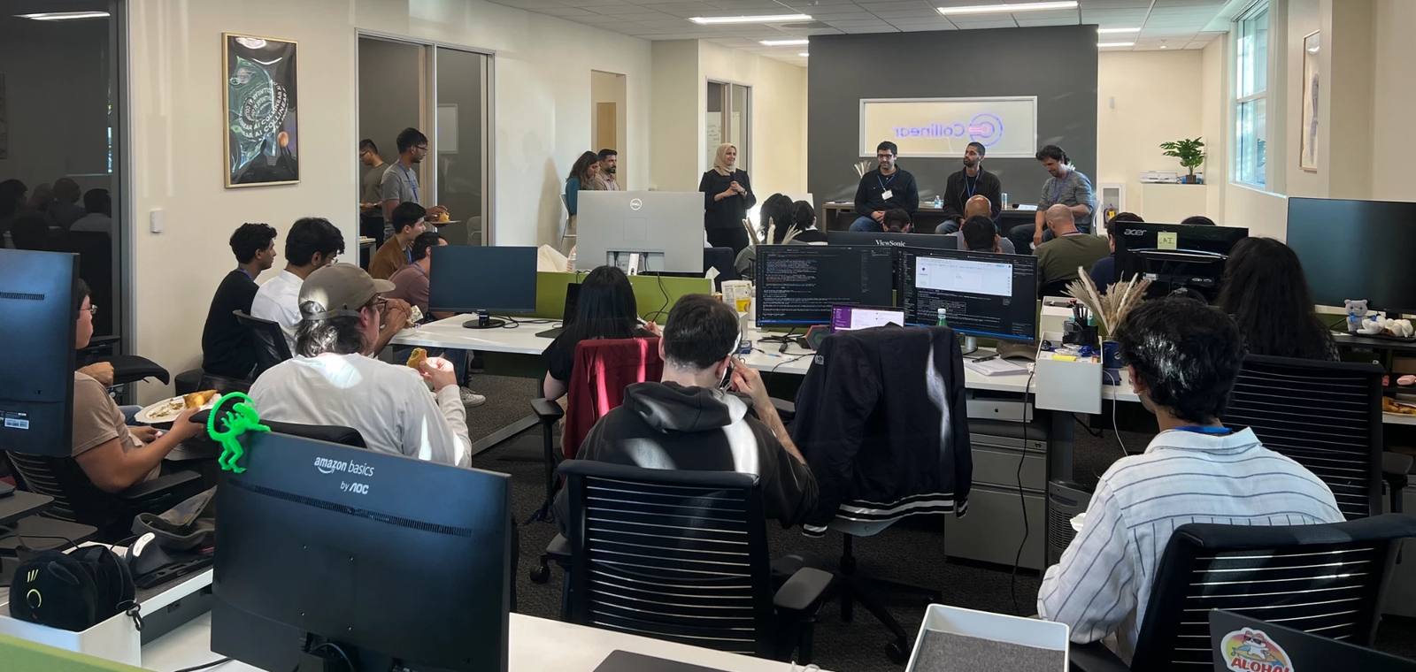 The Collinear team gathered in the Sunnyvale office for an all hands, people at their desks turned toward colleagues presenting beneath the Collinear sign.
