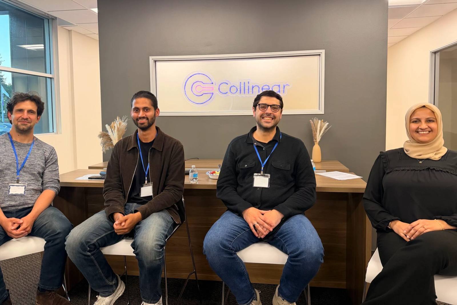 Four Collinear researchers seated together in the office, in front of the lit Collinear sign.