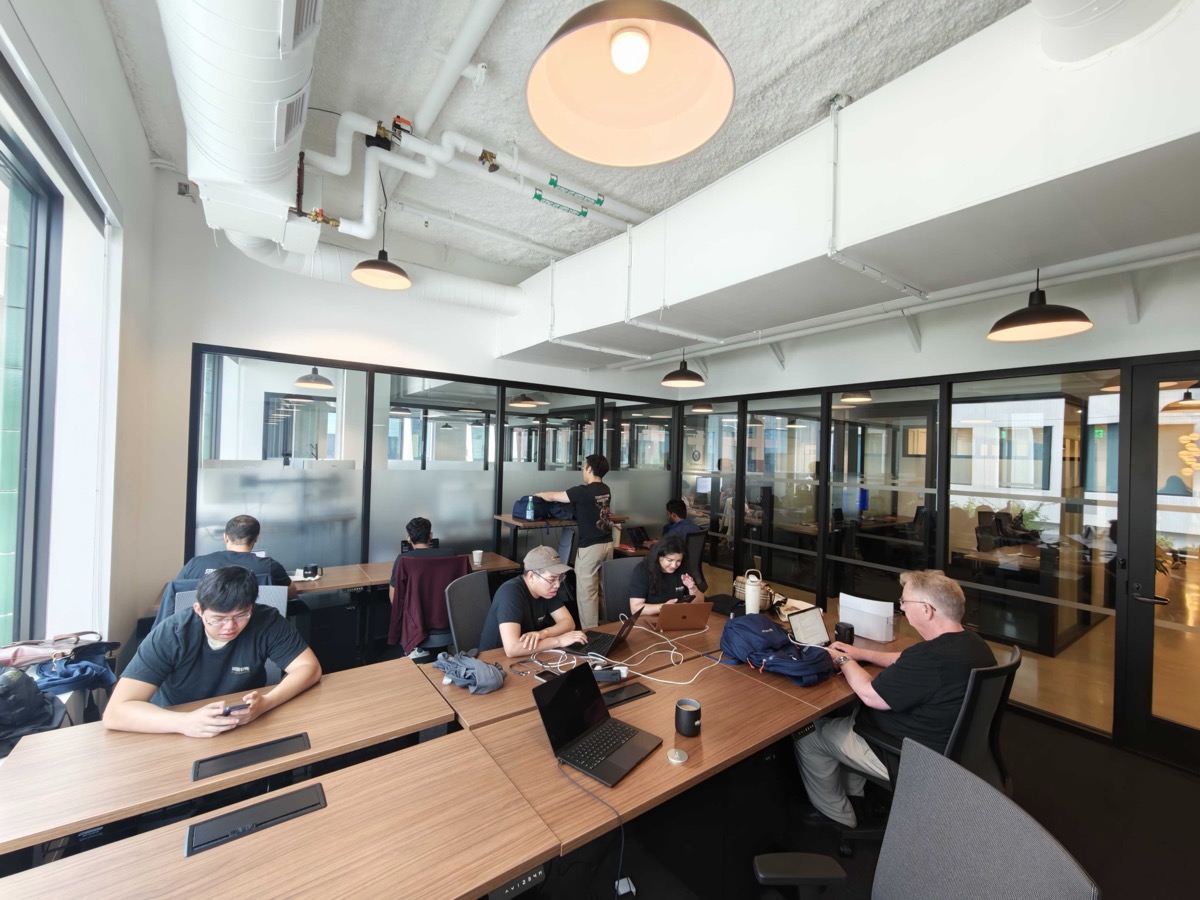 Team members working at shared desks in an open office, with laptops and coffee mugs, one person standing at a height-adjustable desk.