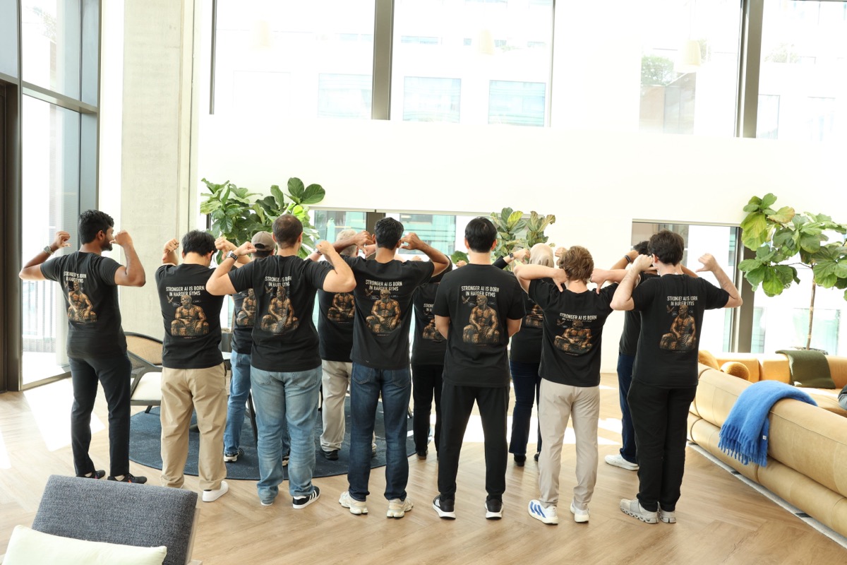 A large group of Collinear team members in matching black 'Harder Gyms' t-shirts, fist-bumping in an office lounge with large windows.