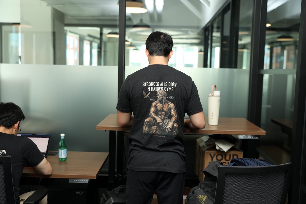 A team member working at a standing desk, back turned to the camera, wearing a 'Stronger AI is born in harder gyms' t-shirt.